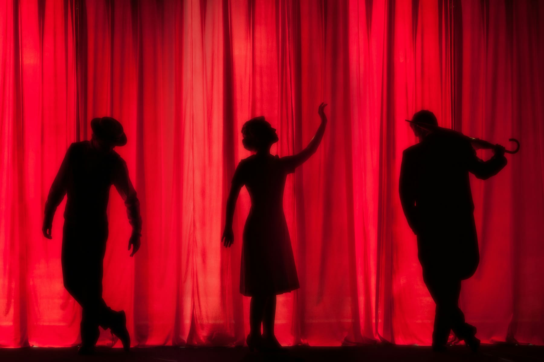 Tre interpreti in silhouette davanti a un sipario rosso durante una rappresentazione teatrale